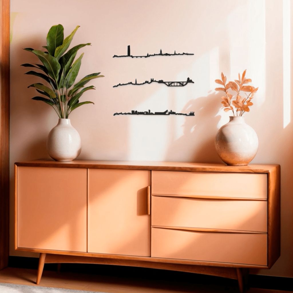 Aparador mid-century de madera y frentes color melocotón con dos jarrones cerámicos —uno con hojas verdes y otro con ramas ocres— sobre un suelo claro; en la pared rosa claro, iluminada por la luz solar, destacan tres siluetas negras de skylines decorativos: Sevilla (arriba), Oporto (centro) y Valencia (abajo).