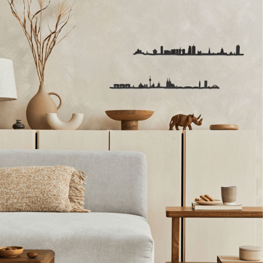 Decoración de salón acogedor con skylines de madera de Köln y Bruselas colgados en la pared, sofá gris claro, muebles y mesas de madera natural, piezas decorativas artesanales y una paleta neutra y cálida.