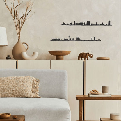 Decoración de salón acogedor con skylines de madera de Köln y Bruselas colgados en la pared, sofá gris claro, muebles y mesas de madera natural, piezas decorativas artesanales y una paleta neutra y cálida.