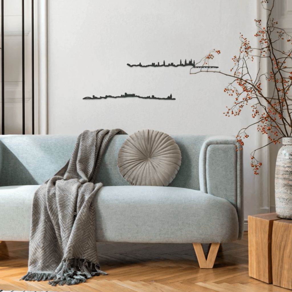 Salón con sofá verde agua, manta gris y cojín circular; suelo de parquet, alfombra de rayas y mesa auxiliar, adornado con skylines de madera de Zaragoza y Huesca en la pared.