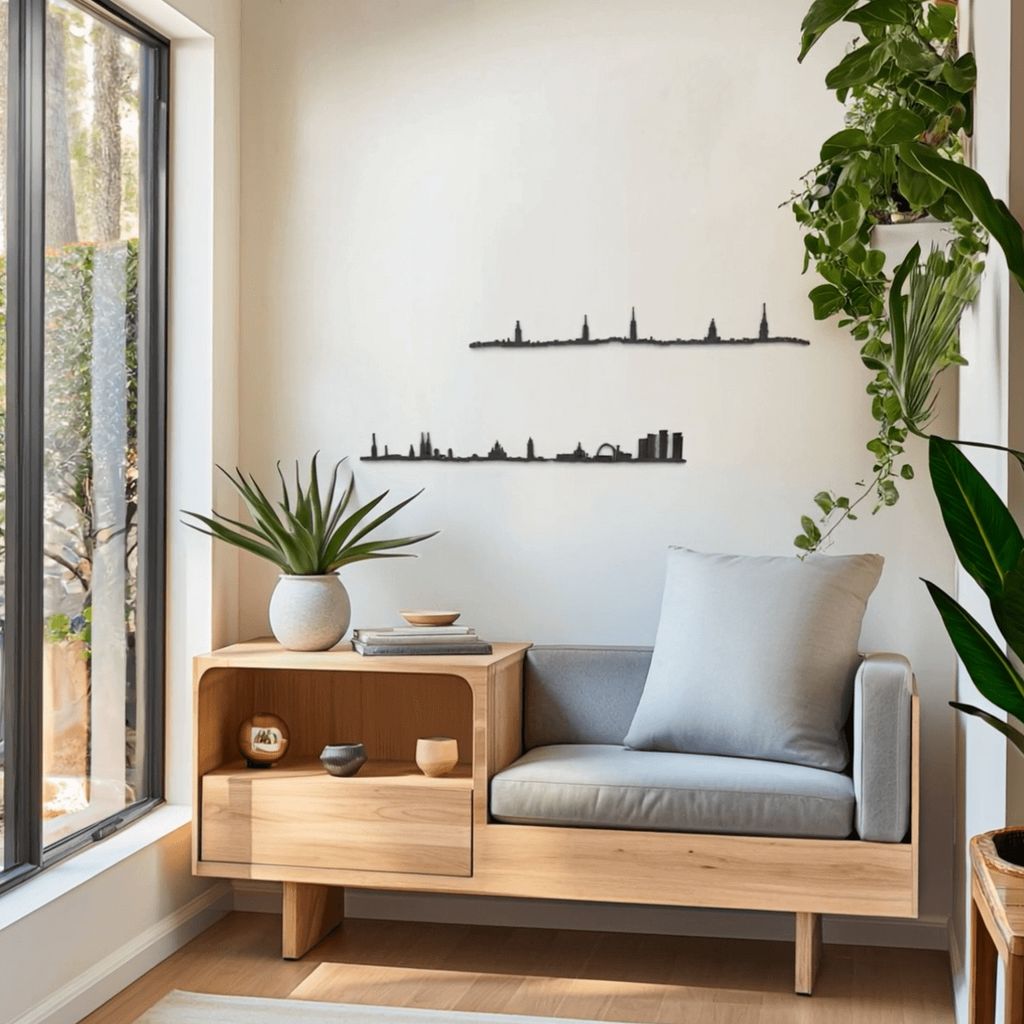 Rincón luminoso junto a un ventanal con banco-mesa de roble claro, cojín gris y maceta de suculenta; a la derecha, planta colgante frondosa. En la pared beige destacan dos siluetas negras de skylines decorativos: Helsinki y Estocolmo (abajo, con el Ayuntamiento y torres del casco antiguo).