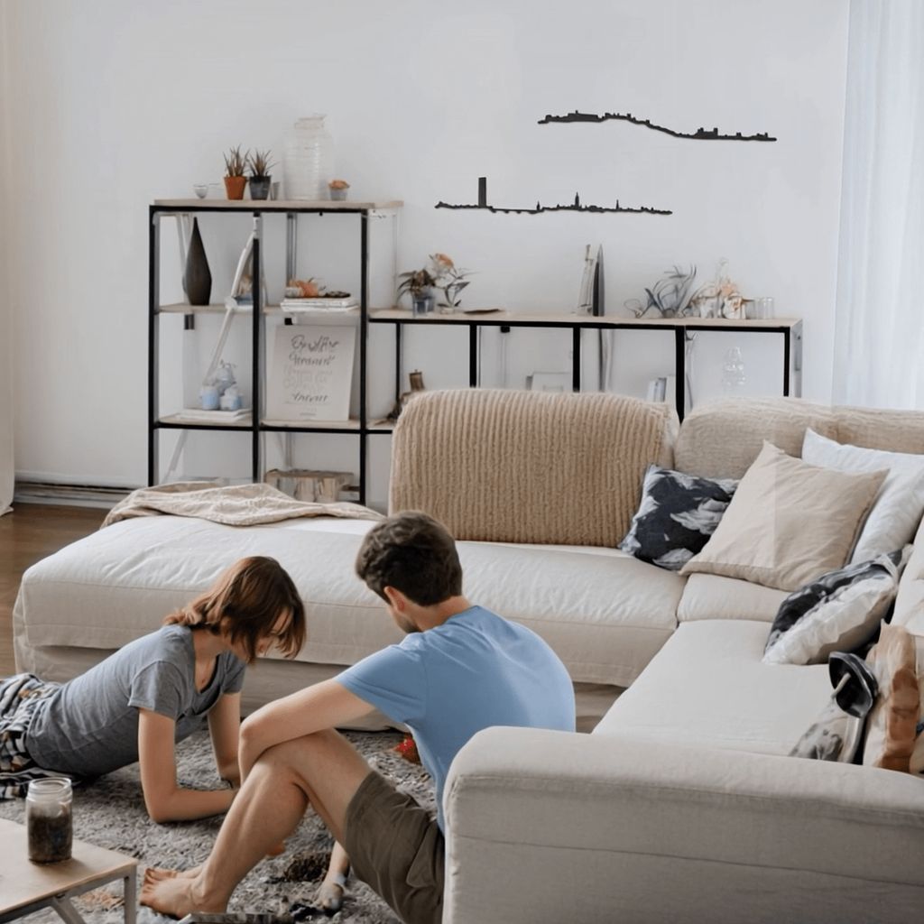 Salón contemporáneo con sofá rinconera beige claro y alfombra gris; una pareja sentada en el suelo juega sobre la alfombra, frente a una mesa auxiliar. Al fondo, estanterías metálicas negras con plantas y objetos decorativos. En la pared blanca se aprecian dos siluetas negras de skylines: Granada (arriba, con la Alhambra sobre la colina) y Sevilla (abajo, con la Torre Sevilla y la Giralda).