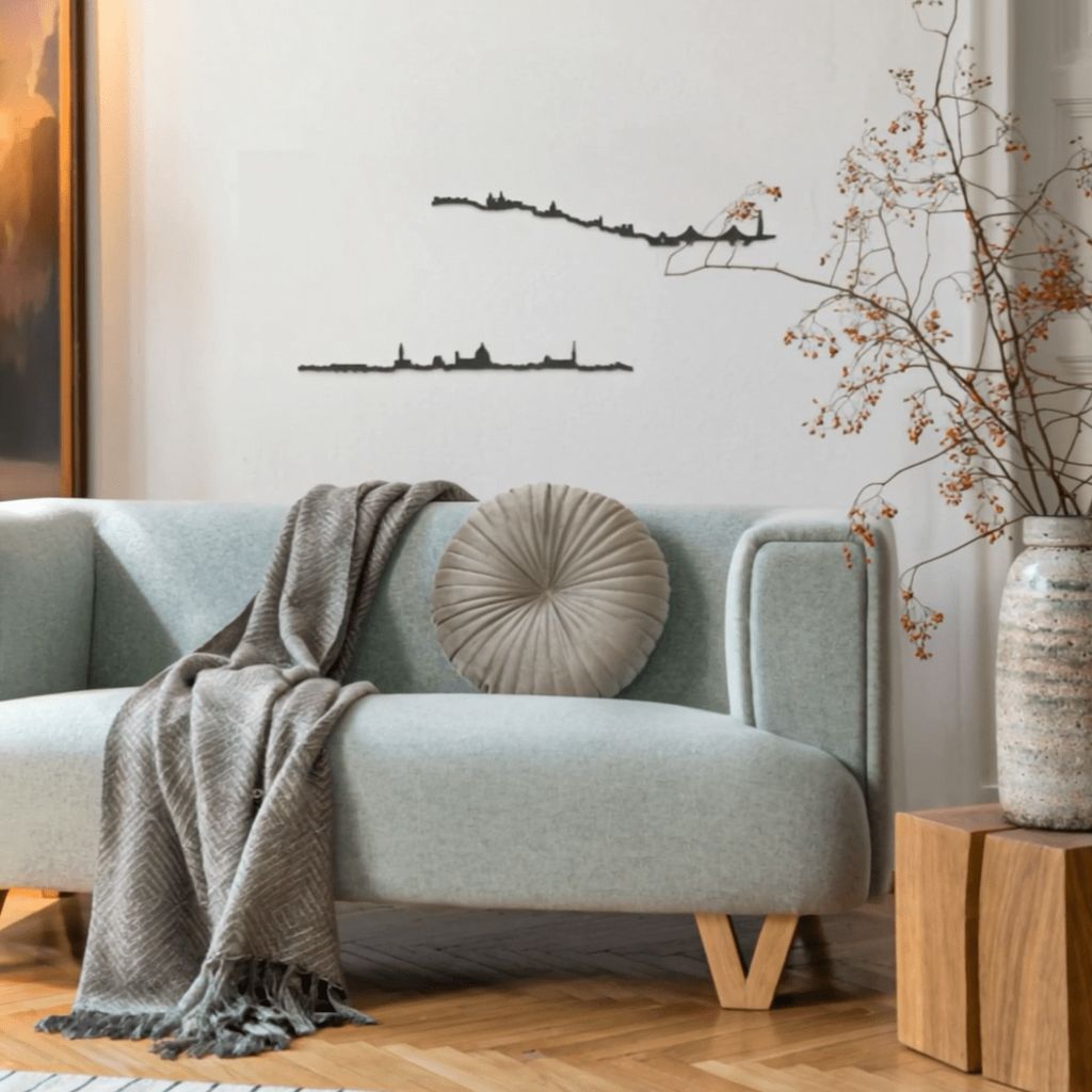 Sala de estar con sofá tapizado en tono menta, cojín redondo beige y manta gris sobre suelo de parquet en espiga; a la derecha, mesa de madera con jarrón esmaltado y ramas con bayas rojas, y a la izquierda se insinúa un cuadro iluminado. En la pared blanca lucen dos siluetas negras de skylines decorativos: Lisboa (arriba, con el puente 25 de Abril) y Florencia (abajo, con la cúpula del Duomo).