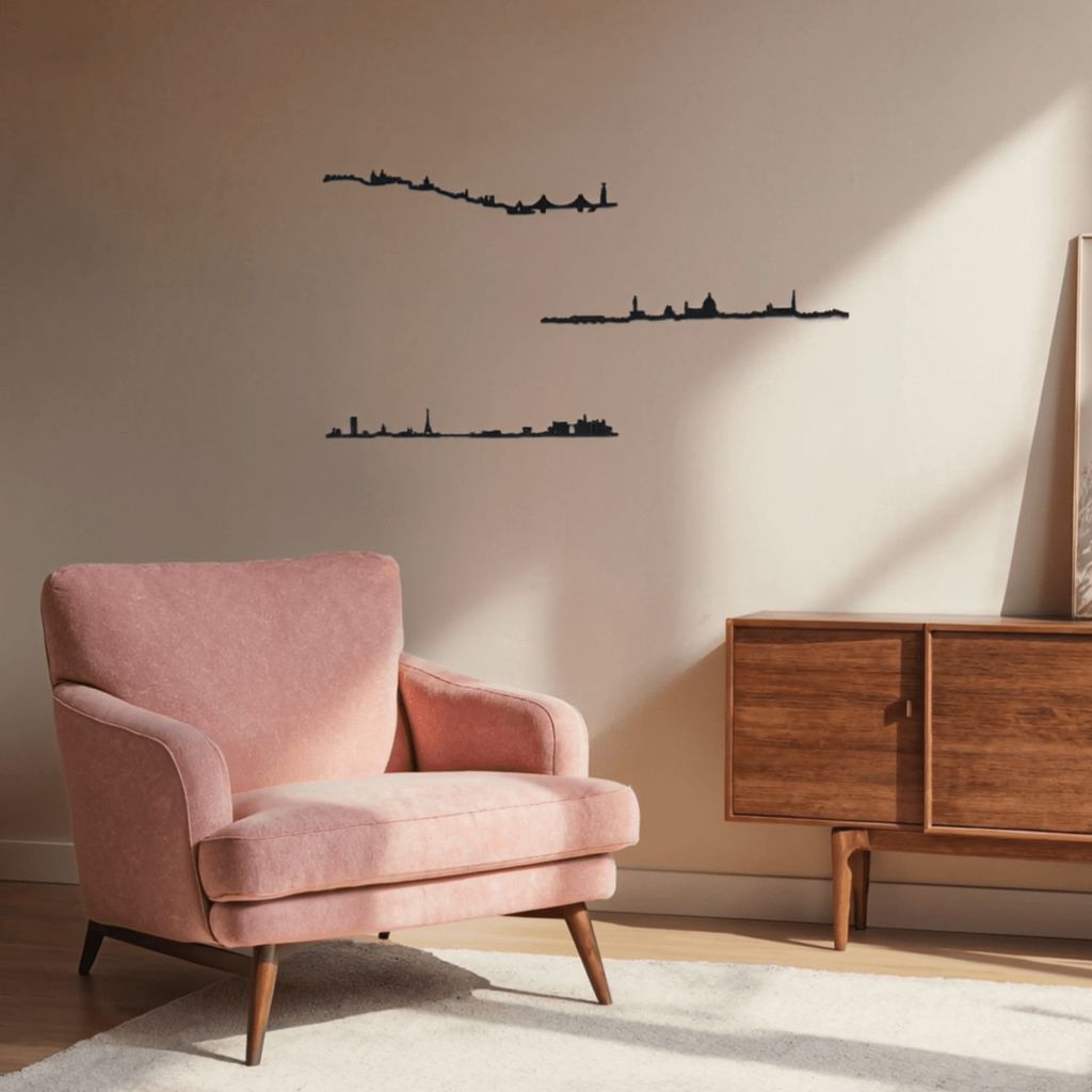 Espacio de lectura de estilo mid-century con butaca rosa empolvada sobre alfombra clara y aparador bajo de madera a la derecha; la pared beige, bañada por luz natural, luce tres siluetas negras de skylines decorativos: París (arriba), Lisboa (centro) y Florencia (abajo).