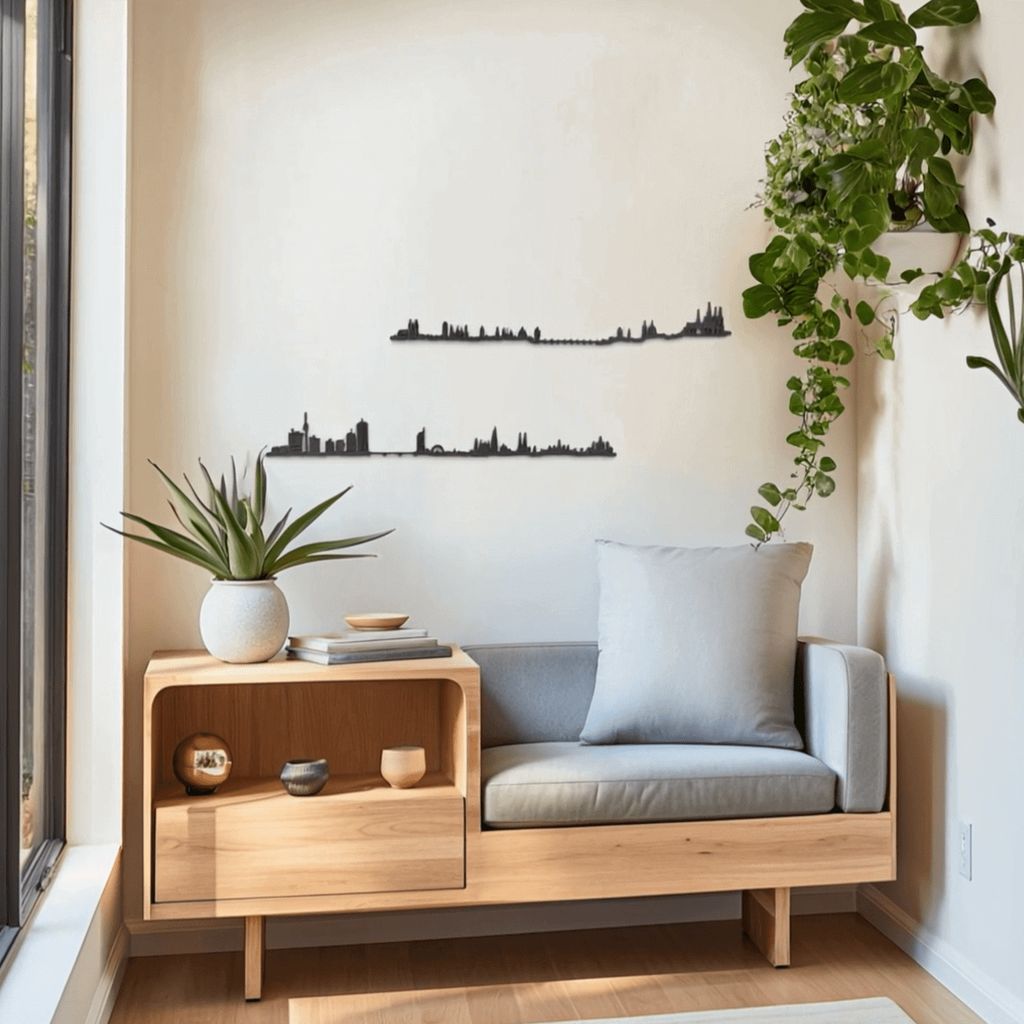 Rincón luminoso con banco-mesa de roble claro y cojín gris, plantas verdes en macetas y gran ventanal a la izquierda; sobre la pared beige se aprecian dos siluetas negras de skylines decorativos: Praga (arriba, con el Puente de Carlos y el castillo) y Viena (abajo, con la catedral de San Esteban y edificios modernos).