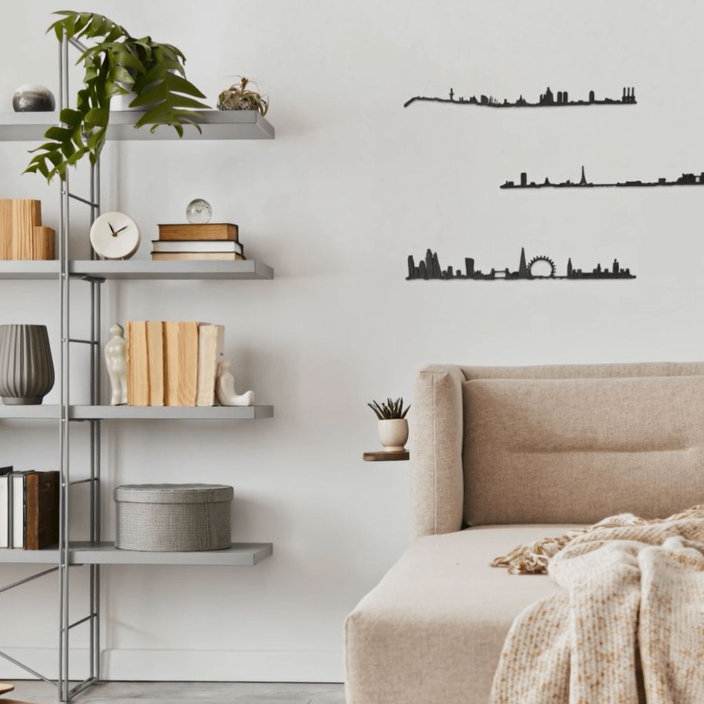 Sala de estar de estilo minimalista con sofá beige y manta jaspeada en primer plano; a la izquierda, estantería metálica gris con libros, reloj, jarrones y una planta colgante. Sobre la pared blanca destacan tres siluetas negras de skylines decorativos: Barcelona (arriba), París (centro) y Londres (abajo).
