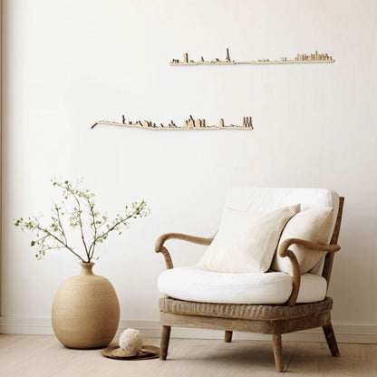 Rincón de lectura minimalista con butaca de ratán y cojines blancos sobre suelo de madera clara; a la izquierda, un jarrón de barro con rama verde y bola decorativa sobre bandeja. En la pared neutra destacan dos siluetas de skylines en madera clara: París (arriba) y Barcelona (abajo).