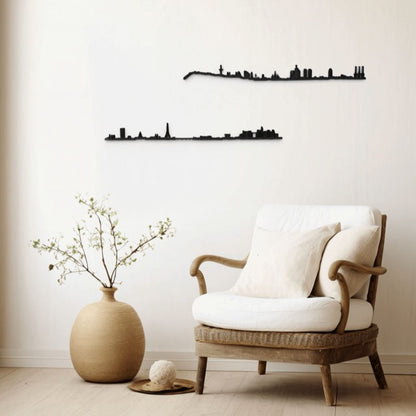 Rincón de lectura minimalista con butaca de ratán y cojines blancos sobre suelo de madera clara; a la izquierda, un jarrón de barro con rama verde y bola decorativa sobre bandeja. En la pared neutra se aprecian dos siluetas negras de skylines: Barcelona (arriba) y París (abajo).