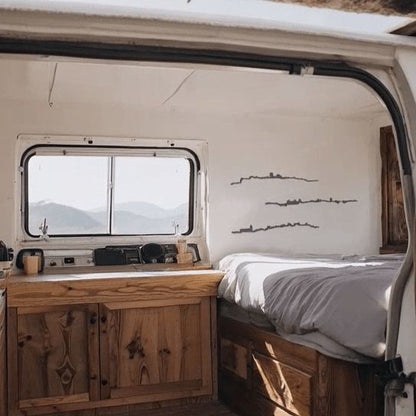 Skylines de Cáceres, Teruel y Zamora en madera negra decorando el interior de una furgoneta camper con muebles de madera natural y cama junto a la ventana con vistas a la montaña.