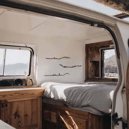 Skylines de Molina de Aragón, Soria y El Burgo de Osma en madera negra decorando el interior de una furgoneta camper con mobiliario rústico de madera, cama junto a la ventana y vistas a las montañas.